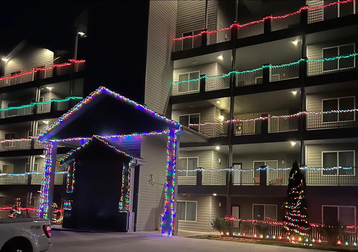The exterior of the property is visible at dusk, with balconies, decorative lights, a prominent entrance awning, and a beautifully lit Christmas tree. The scene is cheerful and designed for celebration, hinting at the vibrant social opportunities available nearby.