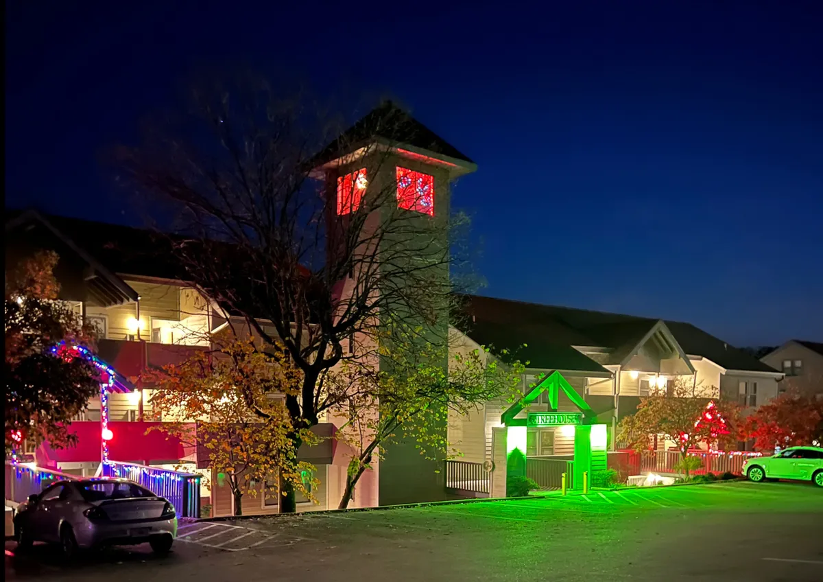 This image shows the exterior of a building adorned with colorful holiday lights, a decorated tree, and an illuminated tower that adds vibrancy to the entrance area. The scene is lively, festive, and invites visitors to explore or gather outside.