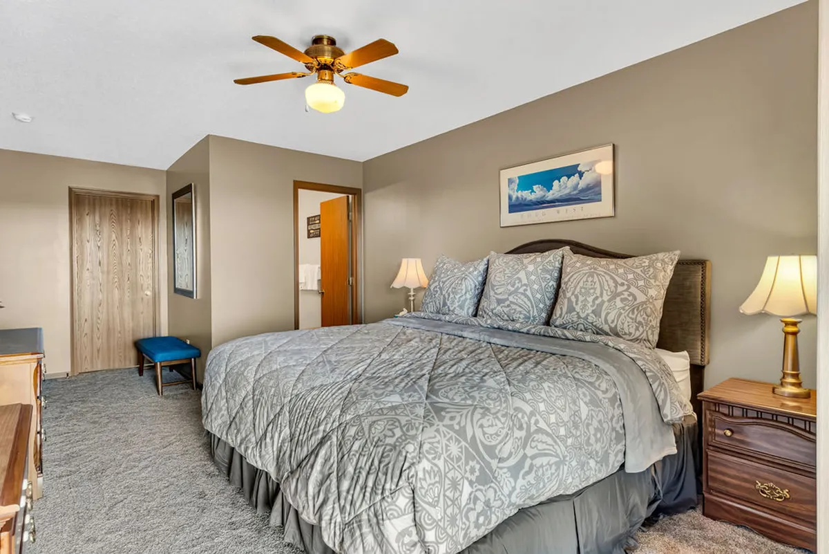 A cozy, inviting bedroom featuring a bed with decorative bedding, nightstands with lamps, a ceiling fan, and tasteful artwork on the wall. Soft natural light filters in, highlighting the serene and relaxing atmosphere tailored for restful nights.
