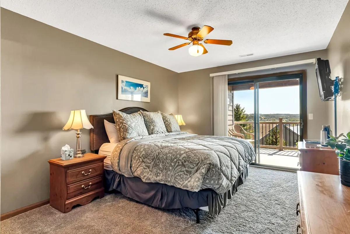A cozy, inviting king bedroom with patterned bedding and a large window offering a scenic view. The positioning of the bed and window suggests direct access to the outdoors, echoing the narrative that the deck is accessible from the king bedroom. Soft natural light adds to the tranquil mood.